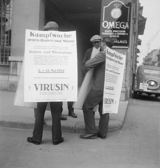 Plakat-Träger mit Plakaten «Kampfwoche gegen Ratten und Mäuse», auf dem Plakat Informationen zur Bekämpfung mit Vertilgungsmittel «Virusin». An der Hausecke Reklameschild für Omega-Uhren, links vergittertes Fenster einer Bank, rechts parkiertes Auto