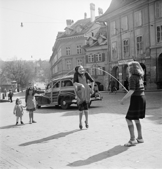 Vier Kinder mit Springseil. Im Hintergrund Nydeggasse und Nydeggbrücke, Auto und Fahrrad am Vorbeifahren, diverse Passanten. Schild von Bäckerei am Haus Gerechtigkeitsgasse Nr. 1