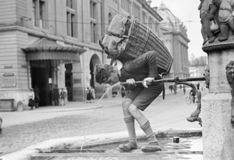 Knabe mit Tragekorb trinkt Wasser am Chindlifresserbrunnen. Im Hintergrund Stadttheater und Kornhaus, links halbrundes Vordach über dem Eingang in den Kornhauskeller