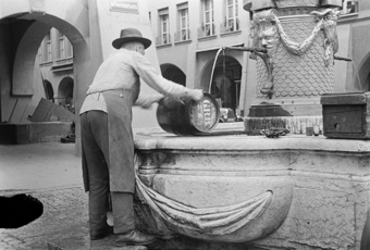 Mann in Arbeitsschürze am Reinigen von Holzfass am Mosesbrunnen, auf dem Brunnenrand Bürste und Holzkiste. Im Hintergrund die Kesslergasse 