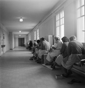 Frauen mit Arbeitsschürzen auf Bänken in einem Korridor sitzend, sich auf Strickarbeit konzentrierend