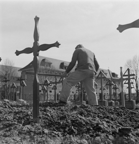 Mann mit Schaufel beim Setzen von Holzkreuzen auf dem Friedhof, im Hintergrund das Hauptgebäude