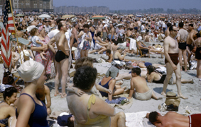 Menschen in Badekleidung dicht gedrängt am Strand stehend, auf Decken und Liegestühlen, einige Sonnenschrime, im Hintergrund Häuser