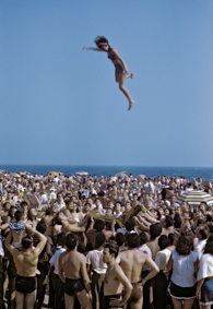 Eine Frau wird mit Hilfe eines Tuchs in die Höhe geworfen und wieder aufgefangen, zahlreiche Badende schauen zu, im Hintergrund das Meer