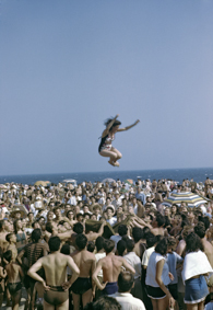 Eine Frau wird mit Hilfe eines Tuchs in die Höhe geworfen und wieder aufgefangen, zahlreiche Badende schauen zu, im Hintergrund das Meer