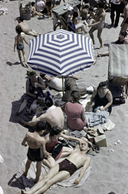 Menschen in Badeanzügen auf Decken und Liegestühlen, unter dem Sonnenschirm eine Familie beim Picknick