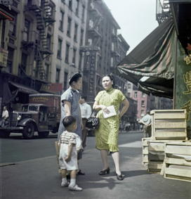Zwei Frauen und ein Kind vor einem Laden in Chinatown, Sonnenstore mit Aufschrift «Chinese Groceries», im Hintergrund Strasse mit Auto und Passanten, Häuserreihe mit Balkonen und Feuerleitern
