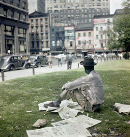 Mann auf dem Rasen eines Parks sitzend, neben ihm  Zeitungsseiten und Papiertüte