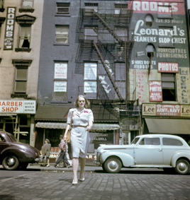 Frau vor einem Gebäude mit Aufschrift «Rooms, Leonard's Seamens Slop Chest» (Zimmer und Laden für Seeleute), im Parterre Barber Shop und Restaurant Schaefer, am Haus Balkone und Feuerleitern, oben links im Bild ein Schild «Rooms Entrance on Water Street»