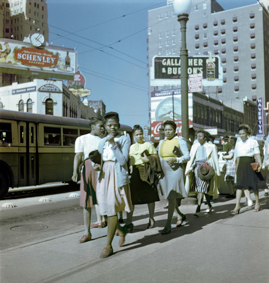 Spazierende Frauen neben einer Strasse mit Bus, im Hintergrund Reklameschilder und Hochhaus, rechts im Bild eine Bus-Haltestelle (Schild Bus Stop «Belmont»)