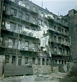 Fünfstöckiges Backsteinhaus mit Balkonen, Holzmast, Wäscheleinen, unten teils kaputte Backsteinmauer mit  Holzbrettern