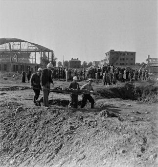 Vier Rotkreuzfreiwillige mit einem exhumierten Leichnam auf einer Tragebahre, im Hintergrund vermutlich Angehörige und Journalisten