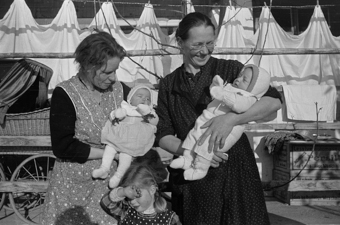 Zwei Frauen mit zwei Babys und einem Mädchen vor der Laube eines Bauernhauses