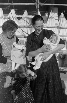 Zwei Frauen mit zwei Babys und einem Mädchen vor der Laube eines Bauernhauses