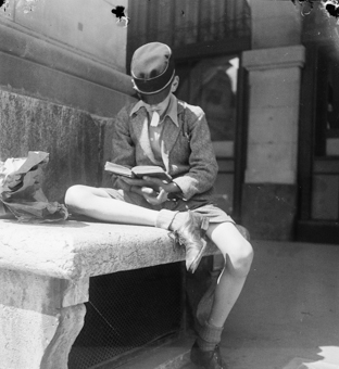 Knabe mit Schülermütze und Buch auf einer Bank sitzend