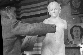Der Künstler hat in der rechten Hand ein Werkzeug, in der linken Hand eine Skizze, an der Wand ein Porträt von Dina Vierny, die ihm während vieler Jahre Modell stand