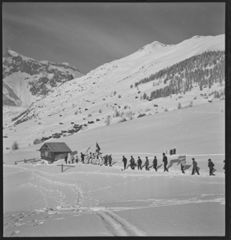 Eine Kolonne von Jungen mit Verkleidungen, Glocken  und Fahnen marschiert durch den Schnee, im Hintergrund verschneite Berglandschaft
