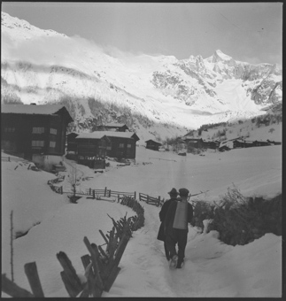 Zwei Männer marschieren durch den Schnee durchs Dorf, dahinter die Bergkulisse
