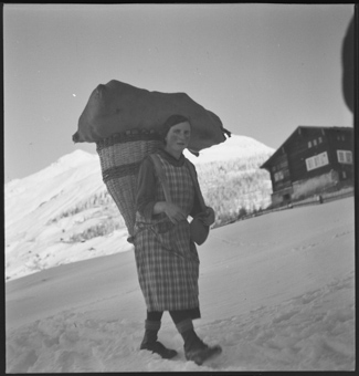 Eine junge Frau schwer beladen mit einem grossen geflochtenen Tragekorb und einem Sack, geht durch den Schnee