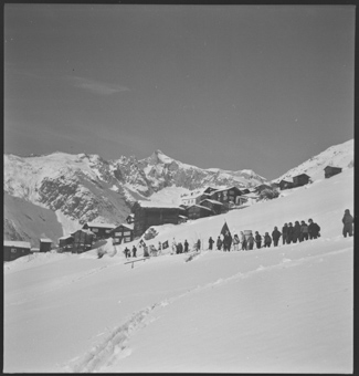 Eine Kolonne von Jungen mit Verkleidungen, Glocken  und Fahnen marschiert durch den Schnee, im Hintergrund das Dorf Bellwald und die verschneite Berglandschaft