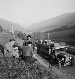 Drei Männer neben voll bepacktem Saurer-Lastwagen, daneben Eisenbahn-Trassee und das Dorf mit Kirche