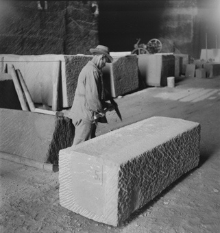 Mann in Arbeitskleidung und Hut bearbeitet einen Steinblock mit dem Pickel, im Hintergrund das Innere des Steinbruchs mit Steinblöcken und Werkzeugen