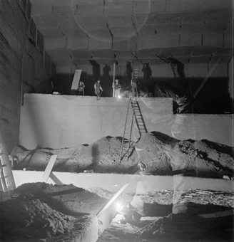 Vier Arbeiter mit Werkzeugen auf Steinwand und auf Leiter, unten mehrere Sandhaufen, rechts im Bild die Umrisse eines weiteren Arbeiters (doppelbelichtete Aufnahme)