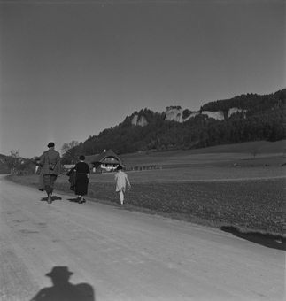 Blick zu den Flühen des Stockern-Steinbruchs, auf der Strasse spaziert eine Familie, im Hintergrund ein Bauernhaus, vorne im Bild der Schatten von Paul Senn