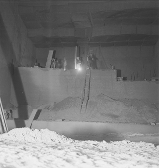 Vier Arbeiter mit Werkzeugen, Leiter und Lampe oben auf einer Steinwand, unten Stein- und Sandhaufen