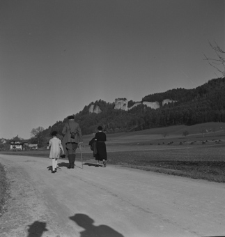 Blick zu den Flühen des Stockern-Steinbruchs, auf der Strasse spaziert eine Familie, im Hintergrund ein Bauernhaus, vorne im Bild der Schatten von Paul Senn und einem Begleiter