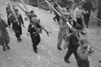 Skifahrer mit geschulterten Skis auf der Dorfstrasse, vorne ein junger Fotograf mit Kamera