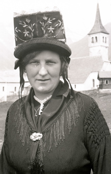Porträt einer Frau mit Tracht und reich verziertem Hut, im Hintergrund die Pfarrkirche von Bellwald
