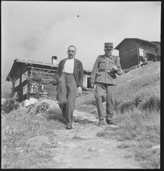 Feldweibel und Gemeindepräsident schreiten auf einem Weg, dahinter Häuser von Bellwald