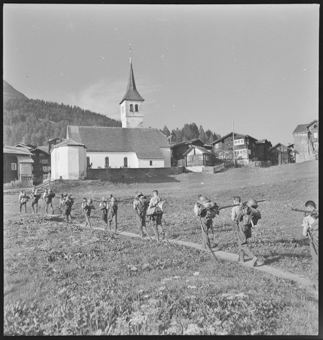 Soldaten mit Gepäck in Einerkolonne zum Dorf marschierend, Kirche und Häuser von Bellwald