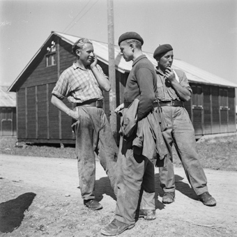 Drei Männer neben den Baracken eines Interniertenlagers